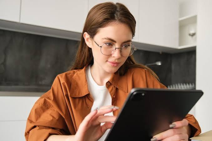 A learner in glasses reading a maths worksheet on a tablet at a kitchen table